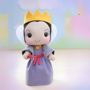 May include: A handmade doll of a character from a fairy tale. The doll has a yellow crown, black hair, and a purple dress. It is holding a red apple and has a white collar. The doll is made of felt and has a smiling expression.