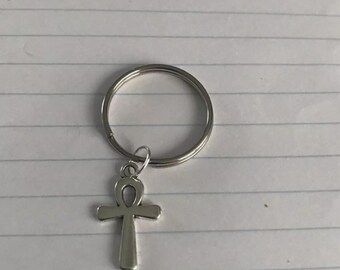 Pode incluir: Um porta-chaves prateado com um pendente do símbolo Ankh. O Ankh, uma cruz com uma alça no topo, está suspenso de um pequeno anel preso a um porta-chaves maior. O porta-chaves é feito de material metálico brilhante.