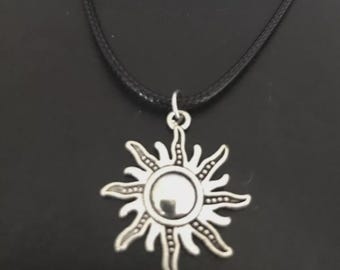 May include: Silver-toned sun pendant necklace with a black cord. The sunburst design features a central, reflective disc surrounded by stylized rays. The necklace is displayed against a dark background, highlighting the pendant's details.