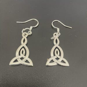 May include: A pair of silver-colored Celtic knot earrings. Each earring features a stylized triquetra design suspended from a simple hook. The earrings are displayed against a dark background.
