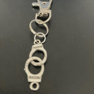 May include: A silver-colored keychain with a lobster clasp, several small rings, and a handcuff charm. The word "FREEDOM" is engraved on the handcuff charm. The keychain is made of metal and is designed to hold keys or other small items.