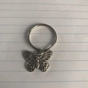 Op de afbeelding: Zilverkleurige vlinder sleutelhanger. De vlinderhanger heeft ingewikkelde uitgesneden details op zijn vleugels en is bevestigd aan een ronde sleutelring. De sleutelhanger is gemaakt van glanzend metaal.