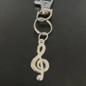 Op de afbeelding: Zilverkleurige sleutelhanger met een G-sleutel bedel. De bedel is bevestigd aan een splitring en een karabijnhaak. De sleutelhanger is ontworpen om aan sleutels of tassen te bevestigen.