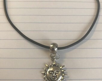 May include: A silver-colored sun pendant necklace with a black cord. The sun pendant has a smiling face and rays extending outward. The necklace is on a lined paper background.