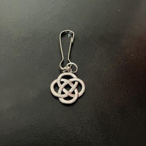Può includere: Un portachiavi color argento con un design a nodo celtico. Il ciondolo è attaccato a un fermaglio di metallo, adatto per chiavi o altri accessori. Il nodo è un anello continuo, che simboleggia l'eternità.
