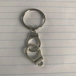 May include: Silver-colored handcuff keychain with the word "FREEDOM" engraved on each cuff. The keychain has a large circular ring for attaching keys. The handcuffs are linked together, and the design is simple and modern.