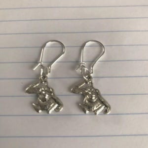 Puede incluir: Un par de pendientes colgantes de color plateado. Cada pendiente presenta un pequeño dije con forma de mono sentado. Los pendientes tienen un cierre de gancho simple.