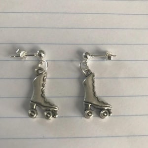 May include: A pair of silver roller skate earrings. Each earring has a detailed roller skate charm with four wheels, suspended from a post and ball stud. The earrings are shown against a lined paper background.