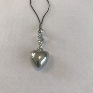 May include: A silver heart charm with a black loop and a silver clasp. The heart is smooth and reflective, hanging from a small metal ring. The charm is likely a pendant or accessory.