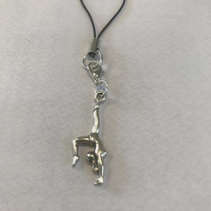 May include: A silver-colored charm featuring a gymnast in a handstand pose. The charm is attached to a black cord with a metal clasp and ring. The gymnast charm is approximately 1 inch in height.