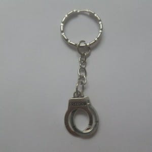 May include: Silver-colored keychain featuring a miniature handcuff charm. The charm has the word "FREEDOM" engraved on it. The keychain includes a split ring and a chain connecting the ring to the handcuff charm.