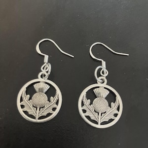 May include: A pair of silver thistle earrings. Each earring has a circular pendant with a thistle design. The design includes the flower and leaves. The earrings have a hook closure.