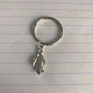 Könnte beinhalten: Ein Schlüsselanhänger mit einem silberfarbenen Pinguin-Anhänger. Der Pinguin-Anhänger ist an einem silberfarbenen Schlüsselring befestigt. Der Schlüsselanhänger befindet sich auf einem linierten Papierhintergrund.