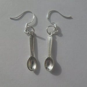 May include: A pair of silver-colored spoon earrings. Each earring features a small spoon charm suspended from a curved ear wire. The spoons have a detailed design and are displayed against a plain white background.