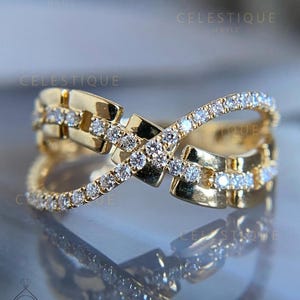 May include: A gold ring with a distinctive crisscross pattern. The ring is adorned with numerous small, glittering white gemstones set along the intersecting bands. The ring has a polished finish, reflecting light. The word "CELESTIQUE" is visible in the image.
