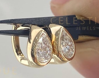 May include: A pair of gold hoop earrings with pear-shaped, clear gemstones. The earrings are held by black tweezers against a light background. The earrings are from Celestique Jewels.