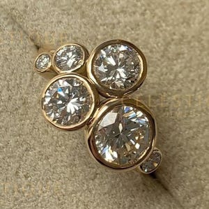 Puede incluir: Un anillo de oro con varias piedras preciosas redondas y transparentes de diferentes tamaños. El anillo está engastado en una banda de oro y se presenta dentro de un joyero beige. La palabra "CELESTIQUE" es visible en el fondo.