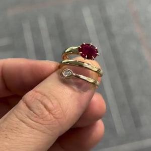Puede incluir: Un anillo de oro tipo bypass con un vibrante rubí rojo de talla ovalada en un extremo y un pequeño diamante blanco en forma de pera en el otro. La banda de oro pulido se envuelve alrededor del dedo, mostrando las formas y colores contrastantes.