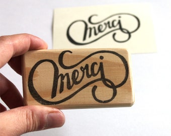May include: A rectangular wooden stamp with the word "Merci" in a stylized, cursive font. The stamp is held in a hand, with a printed version of the design visible in the background. The text is in black against a light wood grain.