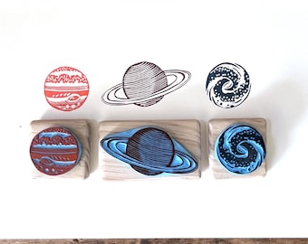 May include: Three rectangular wooden stamps with rubber designs featuring a red planet, a ringed planet, and a spiral galaxy. Above the stamps, corresponding ink impressions are visible on a white surface, showing the detailed celestial patterns.