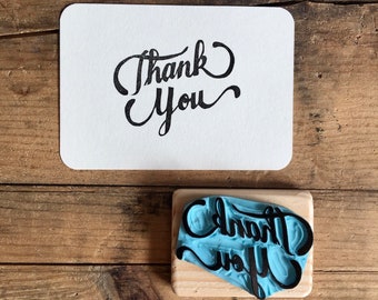 May include: A wooden stamp with a blue rubber surface and the text "Thank You" in black lettering. The stamp is on a wooden base.