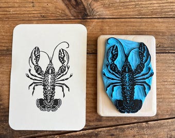 May include: A rectangular rubber stamp with a light blue carved lobster design mounted on a wooden block, positioned next to a white card featuring a black ink print of the same detailed lobster illustration with two large claws and antennae.