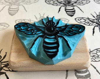 May include: A blue rubber stamp with a detailed black bee design, mounted on a small wooden block. The bee features intricate wing and body details. The background shows bee-themed prints.