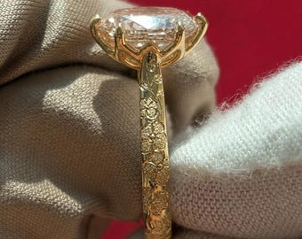 Pode incluir: Um anel de ouro com uma grande pedra preciosa oval, transparente e facetada, presa por uma cravação de múltiplas garras. A banda é feita de ouro amarelo e decorada com um intrincado padrão floral em relevo que cobre toda a superfície.