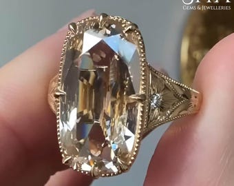 May include: A gold ring featuring a large, elongated, champagne-colored rectangular gemstone with faceted edges. The metal band is intricately engraved with floral patterns and includes a small, round, clear accent stone on each side.