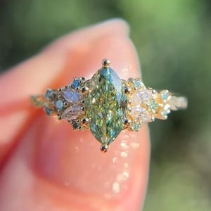 May include: A gold ring featuring a central marquise-cut green gemstone flanked by smaller marquise and round-cut white stones. The band is thin and metallic. The Siya Gems & Jewelleries logo is visible in the top right corner of the image.