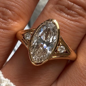 Peut inclure: Une bague en or avec un grand diamant central taille ovale serti dans une monture lisse. L'anneau comprend deux petits diamants ronds d'accent, un de chaque côté de la pierre centrale, intégrés dans le corps de bague en or jaune poli.