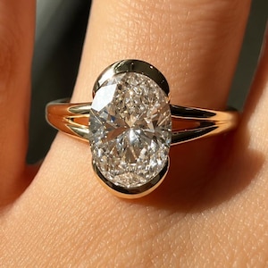 Puede incluir: Un anillo de oro con una gran piedra preciosa transparente de talla ovalada y facetada, montada en un engaste de bisel liso. La banda del anillo se divide en dos segmentos finos de oro pulido que se unen en la base del engaste central.