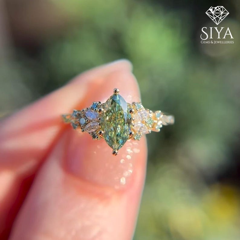 May include: A gold ring featuring a large, marquise-cut, light green gemstone as the centerpiece. The ring is adorned with smaller, round and pear-shaped gemstones in shades of white and blue. The SIYA Gems & Jewelleries logo is visible in the upper right corner.