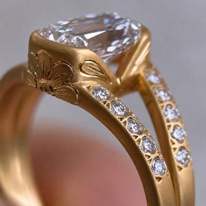 Peut inclure: Une bague en or avec une grande pierre transparente à facettes rectangulaires montée sur un anneau en or brossé. Le sertissage est orné d'un motif floral gravé sur le côté, et la tige fendue est accentuée de petites pierres rondes.
