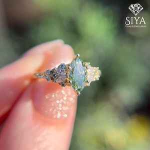 May include: A gold ring featuring a central marquise-cut light green gemstone flanked by smaller marquise and round-cut clear stones on each side. The band is thin and metallic. The text SIYA GEMS & JEWELLERIES is visible in the corner.