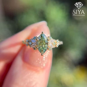 May include: A gold ring featuring a large, marquise-cut, light green gemstone as the focal point. The ring is adorned with smaller, multi-colored gemstones, including white, blue, and green, creating a cluster design. The ring is set against a blurred green background.