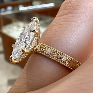 May include: A yellow gold ring featuring a large marquise-cut center stone held by six prongs. The band is decorated with intricate engraved scrollwork patterns and milgrain edges, with small accent stones set along the basket of the setting.