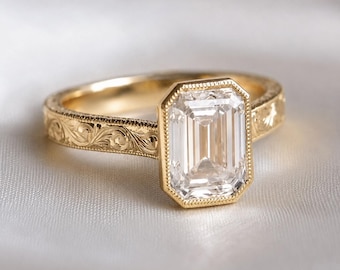 May include: A gold ring featuring a large, clear, emerald-cut gemstone set in a bezel with beaded edges. The band is crafted from yellow gold and displays intricate, engraved scrollwork patterns along the entire outer surface of the shank.