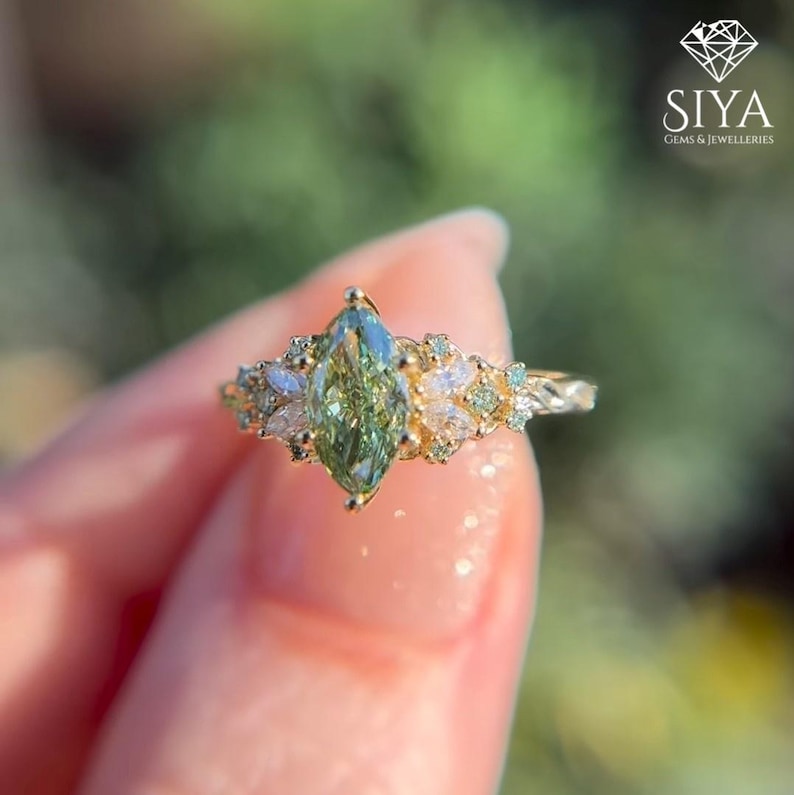 May include: A gold ring featuring a marquise-cut, light green gemstone as the centerpiece, surrounded by smaller white and green stones. The ring has a delicate, detailed band. The SIYA Gems & Jewelleries logo is in the upper right corner.