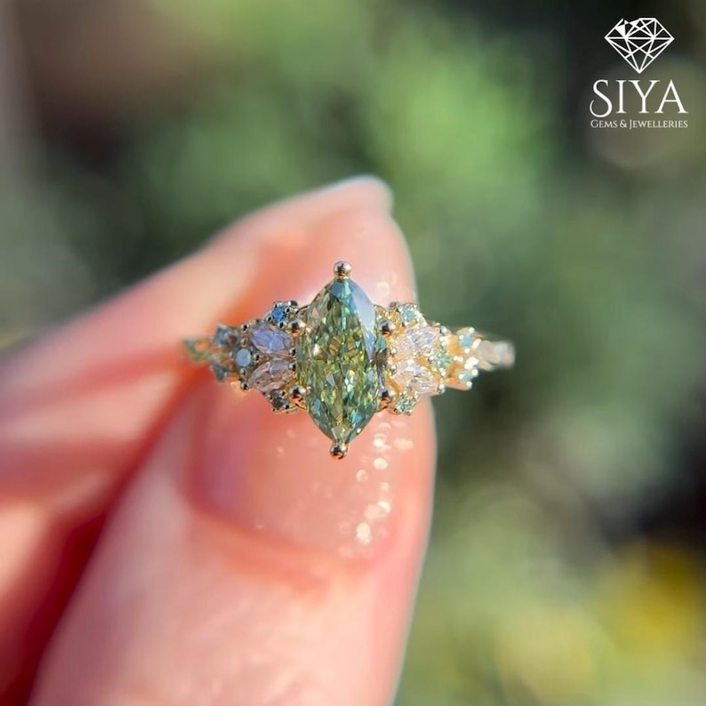 May include: A gold ring featuring a central marquise-cut green gemstone flanked by smaller marquise and round-cut white stones. The band is thin and metallic. The Siya Gems & Jewelleries logo is visible in the top right corner of the image.