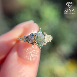 May include: A gold ring featuring a central marquise-cut light green gemstone flanked by smaller marquise and round-cut clear stones. The band has a textured, branch-like design. The logo SIYA JEWELLERIES is visible in the corner.