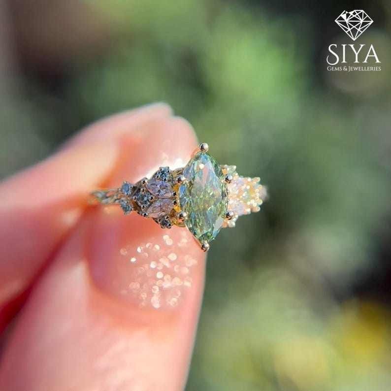 May include: A gold ring featuring a central marquise-cut light green gemstone flanked by smaller marquise and round-cut clear stones. The band has a textured, branch-like design. The logo SIYA JEWELLERIES is visible in the corner.