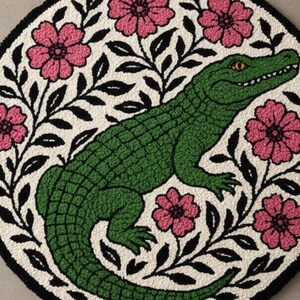 以下が含まれることがあります： 中央に緑のアリゲーター、オフホワイトの背景にピンクの花と黒い葉が描かれた丸いラグ。ラグは黒い縁で囲まれ、装飾的で気まぐれなデザインになっています。