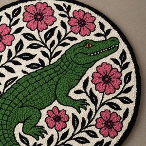 以下が含まれることがあります： クリーム色の背景に、緑のアリゲーターのデザイン、ピンクの花、黒い葉が描かれた丸いラグ。 ラグには黒い縁取りがあります。 このデザインは、自然とアートを組み合わせた遊び心のあるもので、柔らかく質感のある素材で作られている可能性があります。