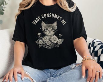 May include: Black t-shirt featuring a cat illustration surrounded by daisies. The text "RAGE CONSUMES ME" is arched above the cat's head. The shirt is worn by a person wearing blue jeans.