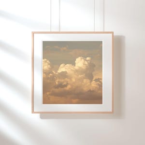 May include: A framed print featuring a photograph of a cloudscape. The image showcases fluffy, white clouds against a backdrop of a light blue sky. The frame is a light wood color, and the print is displayed on a white wall.