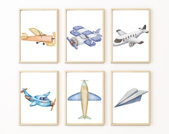 May include: Set of six framed watercolor prints featuring various airplanes. The prints showcase different aircraft designs in shades of orange, blue, and white, including a paper airplane. The artwork is set against a white background.