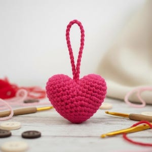 May include: A vibrant pink crocheted heart with a hanging loop. The image features the heart as the central element, with yarn, crochet hooks, and buttons scattered around, indicating a crafting project. The heart's texture is prominent.