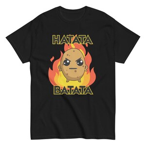 以下が含まれることがあります： 黒いTシャツ。 炎に囲まれた漫画のジャガイモのキャラクターが描かれています。 「HATATA BATATA」の文字がジャガイモの上と下に黄色でプリントされています。 ジャガイモは怒った表情をしています。 デザインはTシャツの中央に配置されています。