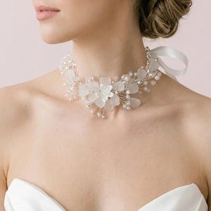 Puede incluir: Un collar de gargantilla nupcial floral blanco. El collar presenta delicadas flores blancas, pequeñas perlas y detalles de alambre plateado. Una cinta de satén blanco asegura el collar en la parte posterior. La gargantilla está diseñada para una boda u ocasión especial.